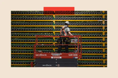 LARS HAGBERG/AFP via Getty A man in a safety helmet stands on a platform in front of a bank of Bitcoin mining computers