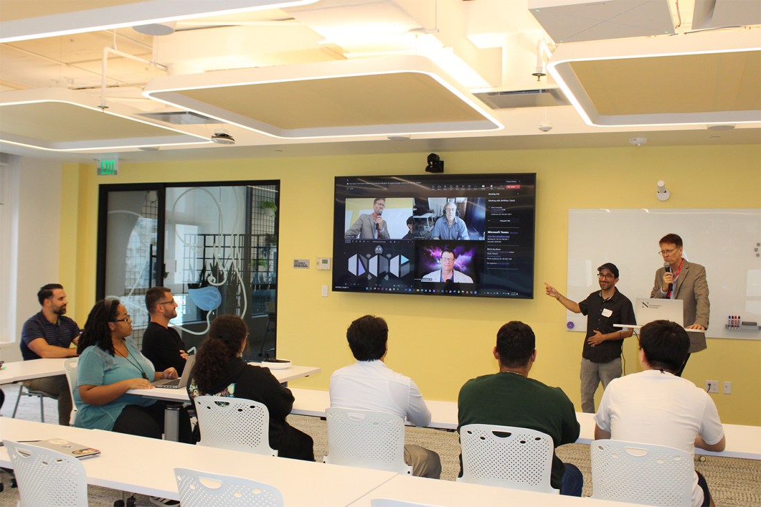 Two people standing at the front of the room next to a TV. A group of other people are sitting in tables around the TV.