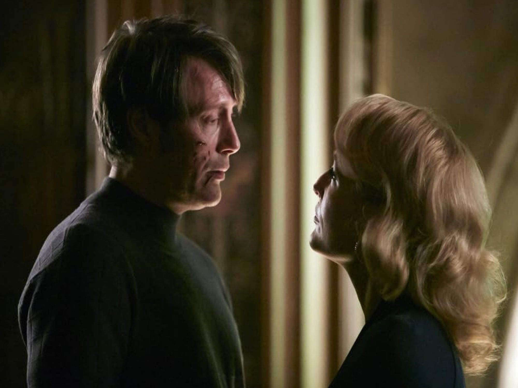 Mads Mikkelsen as Hannibal Lecter. He has cuts on his face and wears a dark turtleneck. Opposite him is Gillian Anderson as Bedelia du Maurier. She stares up at him, also decked in black.