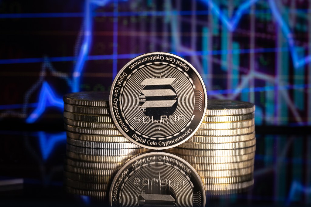 A Solana (SOL) cryptocurrency coin is placed on a stack of coins with blurred price charts in the background.