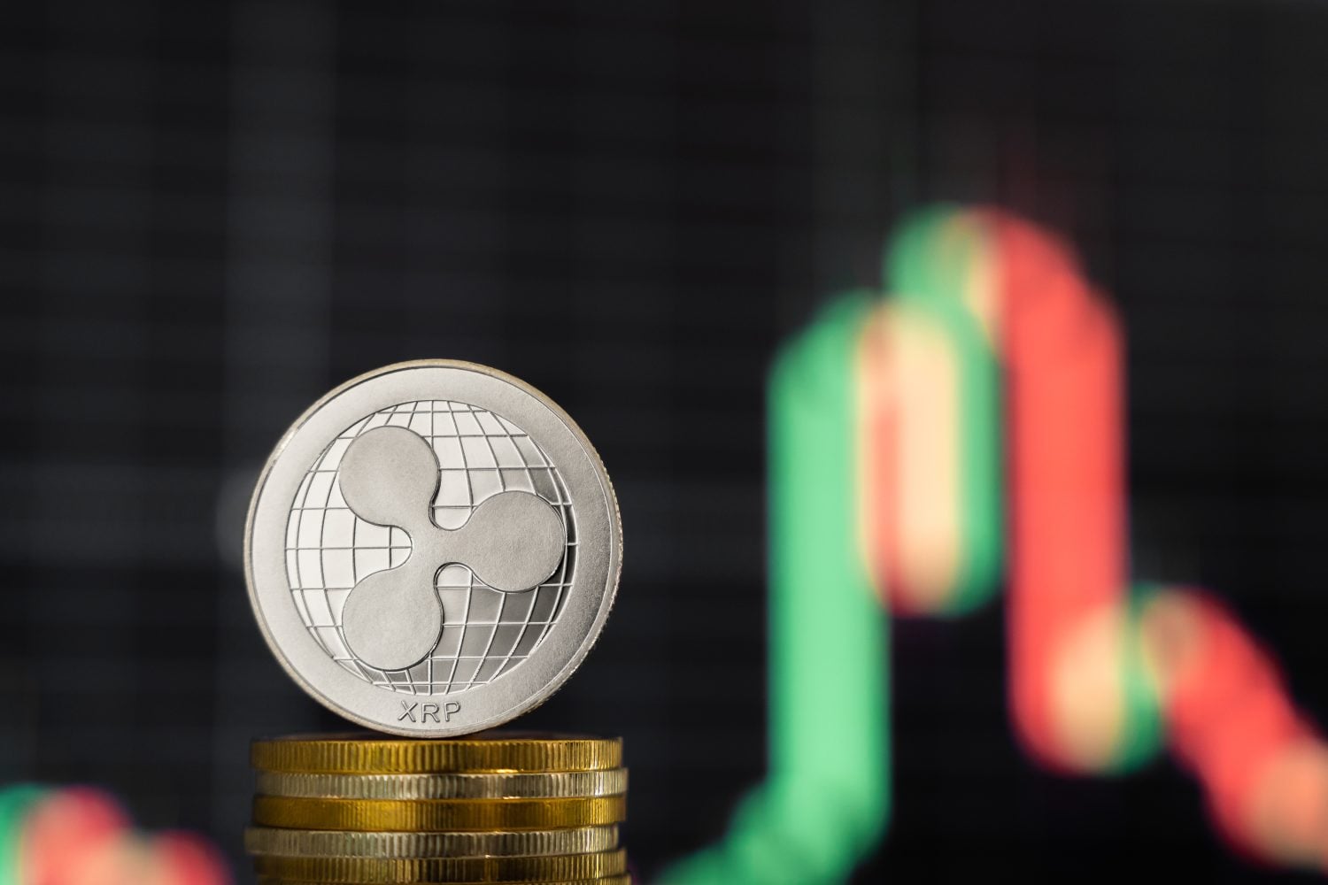 A silver XRP cryptocurrency coin stands upright on a stack of several gold coins. The XRP coin features its distinct logo over a grid-like globe design. The background is a dark, blurred financial chart with prominent wavy lines of red and green, suggesting market movements.